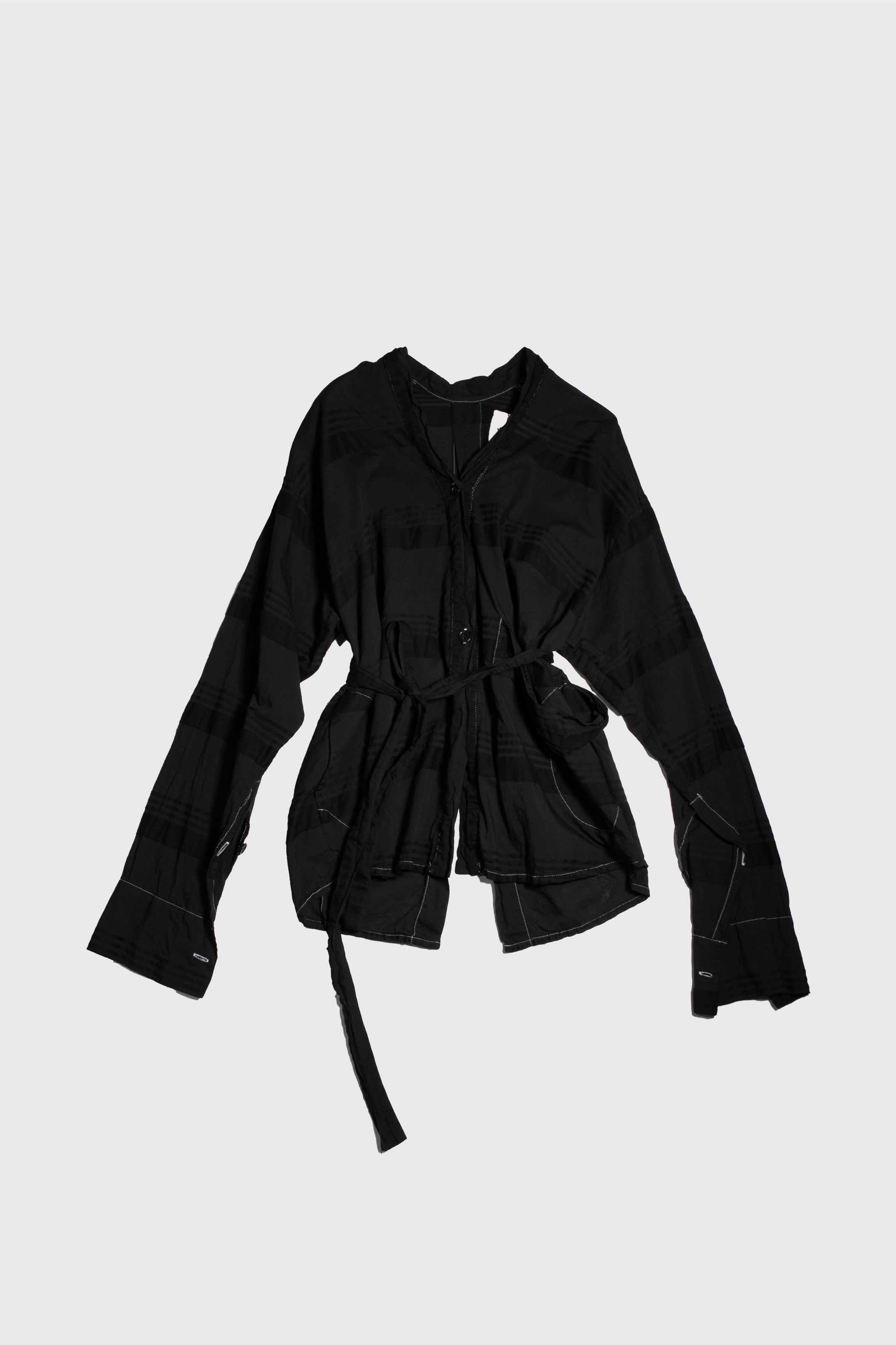 Black cotton shirt laid flat showing angled V-neck collar, front button closure, and self-tie waist belt