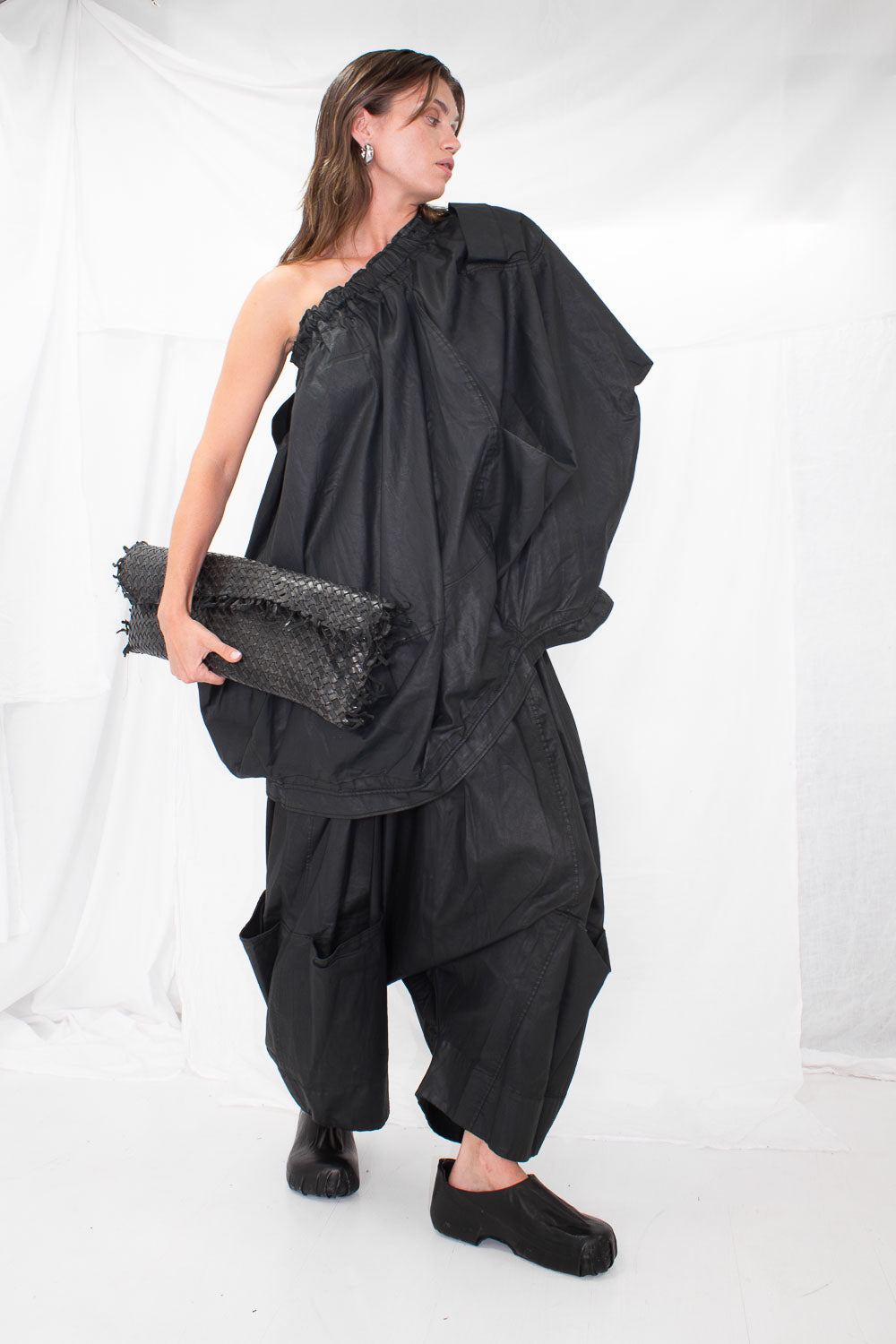 Model wearing black oversized top styled as one-shoulder cape with matching clutch and shoes