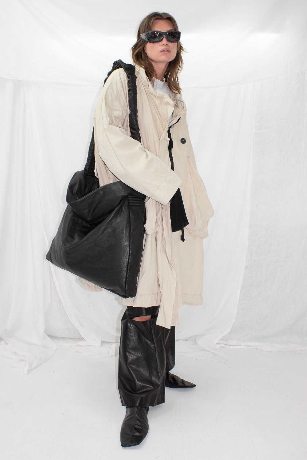 Model wearing oversized black leather bag with beige coat in studio setting