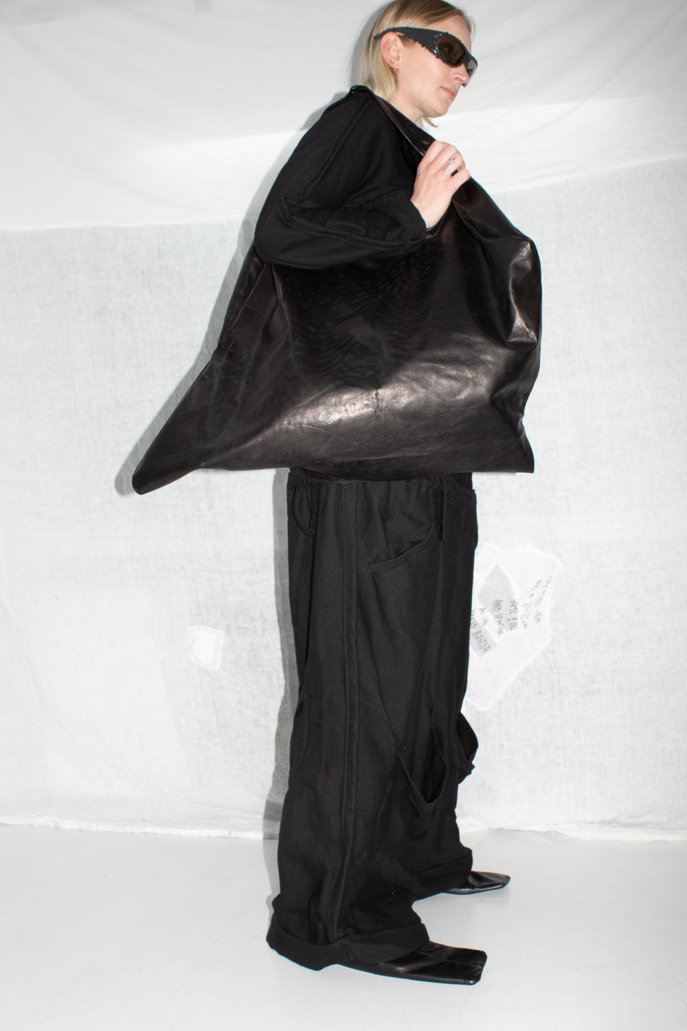 Model in sunglasses holding large leather bag, wearing black trousers with visible waistband styling