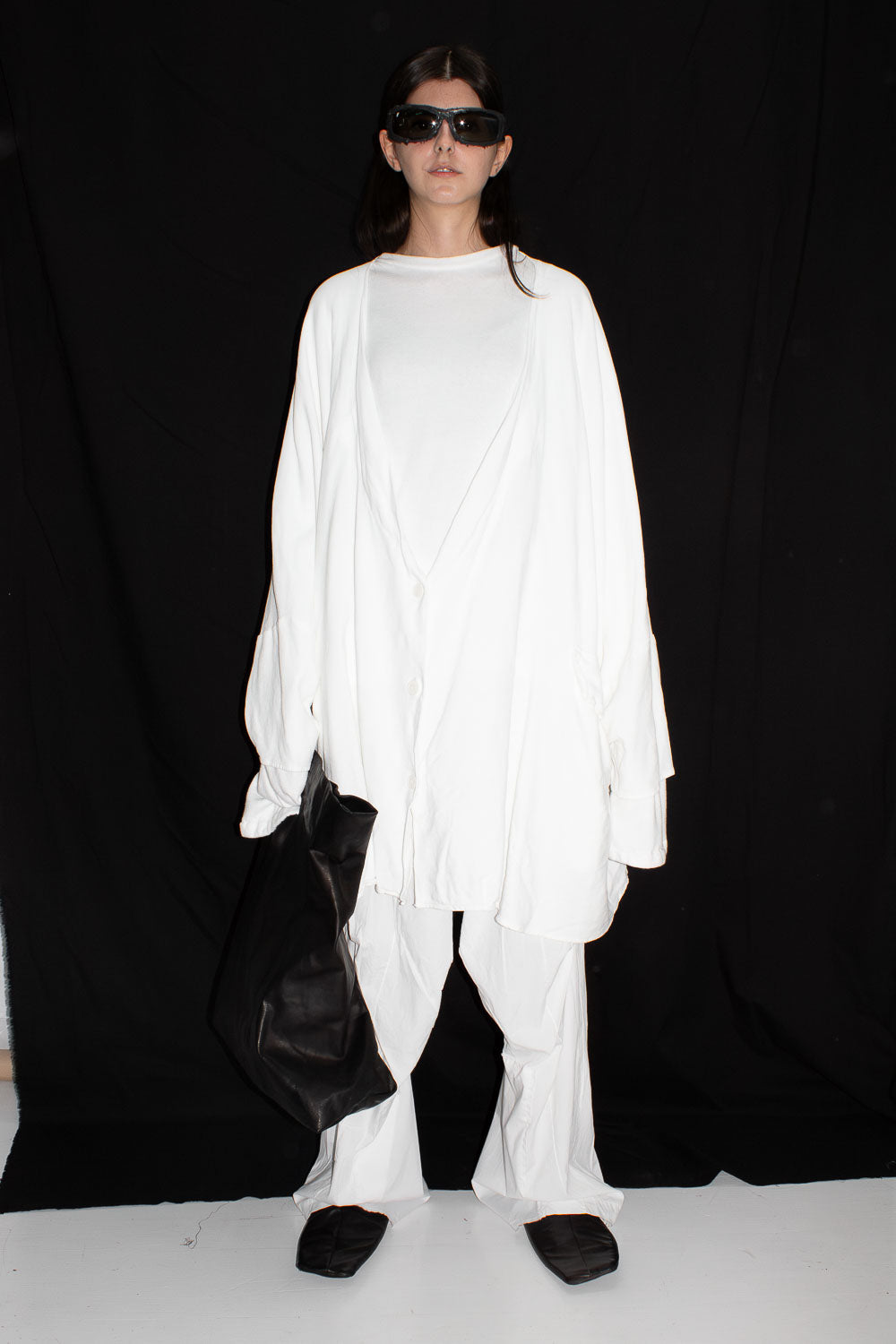 Model wearing white oversized cardigan with black sunglasses and black bag, full body front view