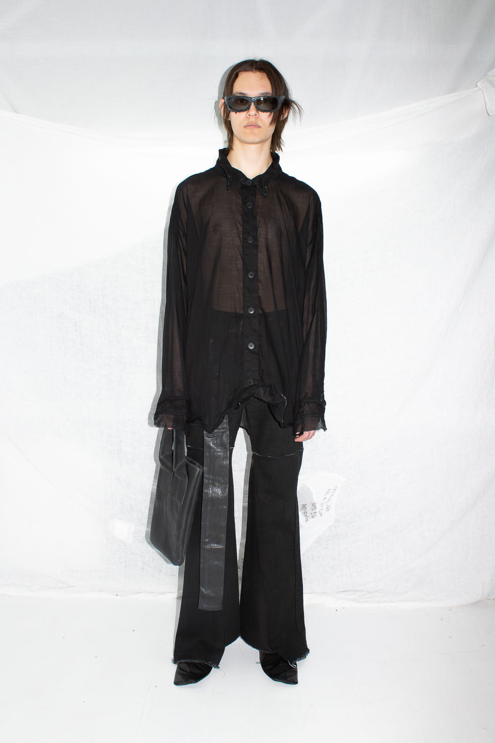 Front view of model in sunglasses wearing black sheer shirt and wide trousers with leather tote