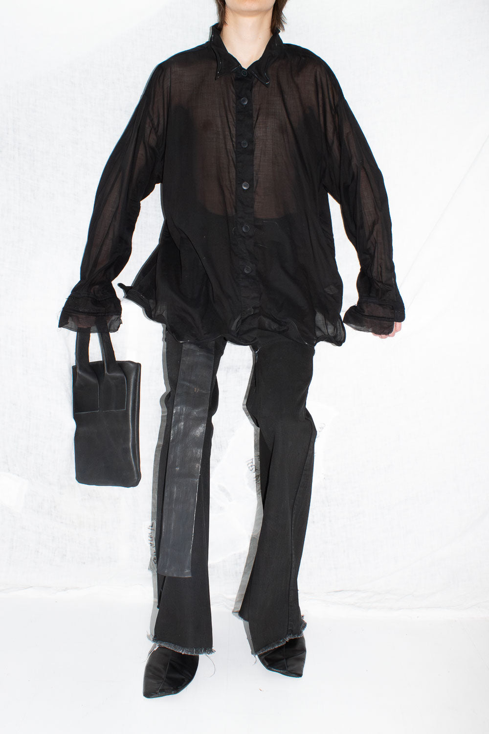 Model wearing oversized black sheer shirt and trousers holding black leather tote