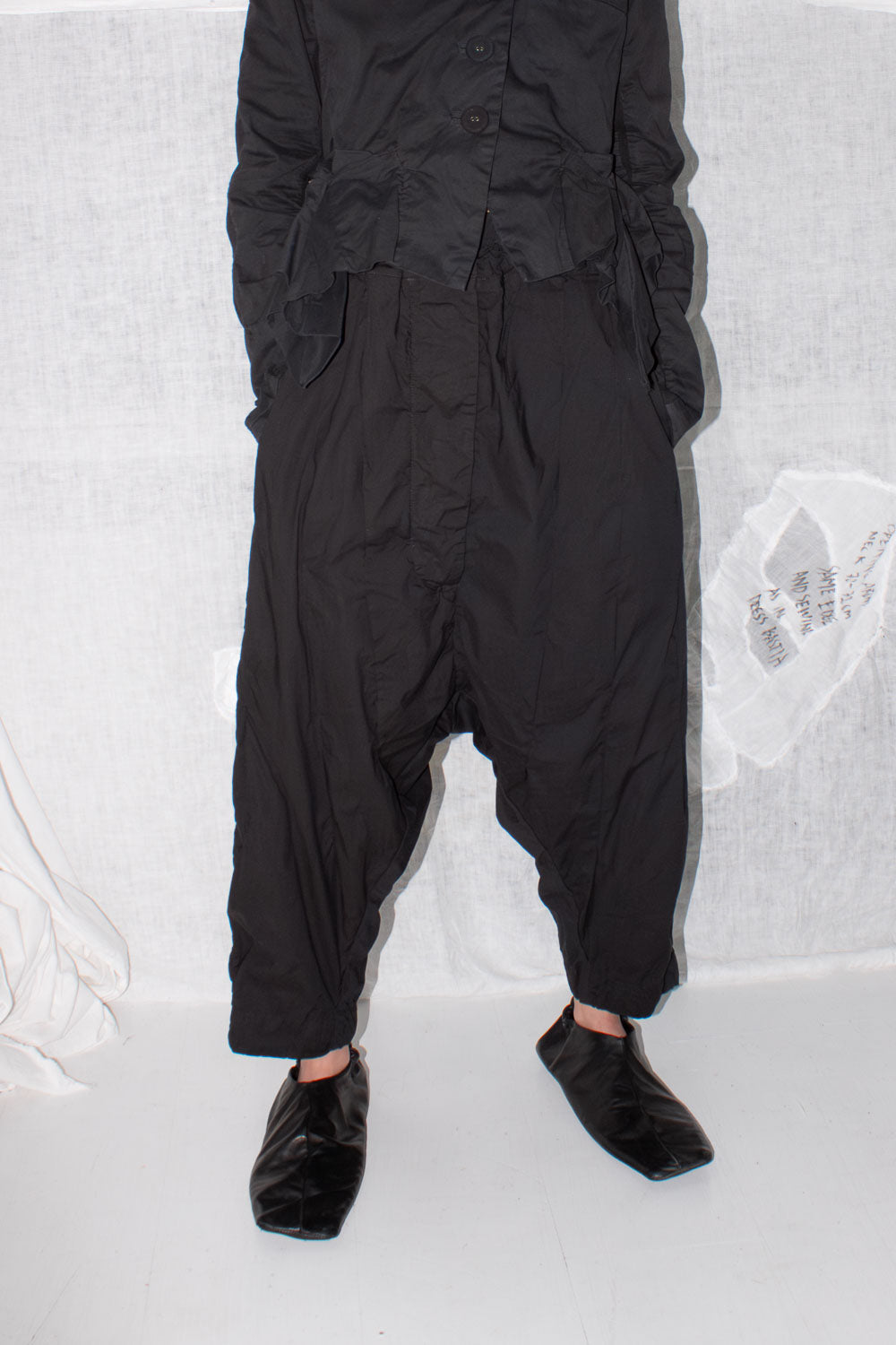 Black pleated trousers worn with leather shoes, showing dropped crotch silhouette and cropped length