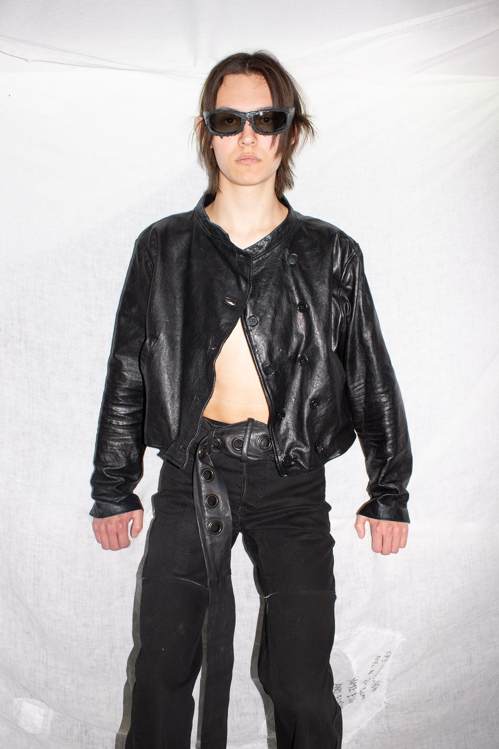 Model in black leather cropped jacket buttoned asymmetrically with structured hem, paired with black leather pants