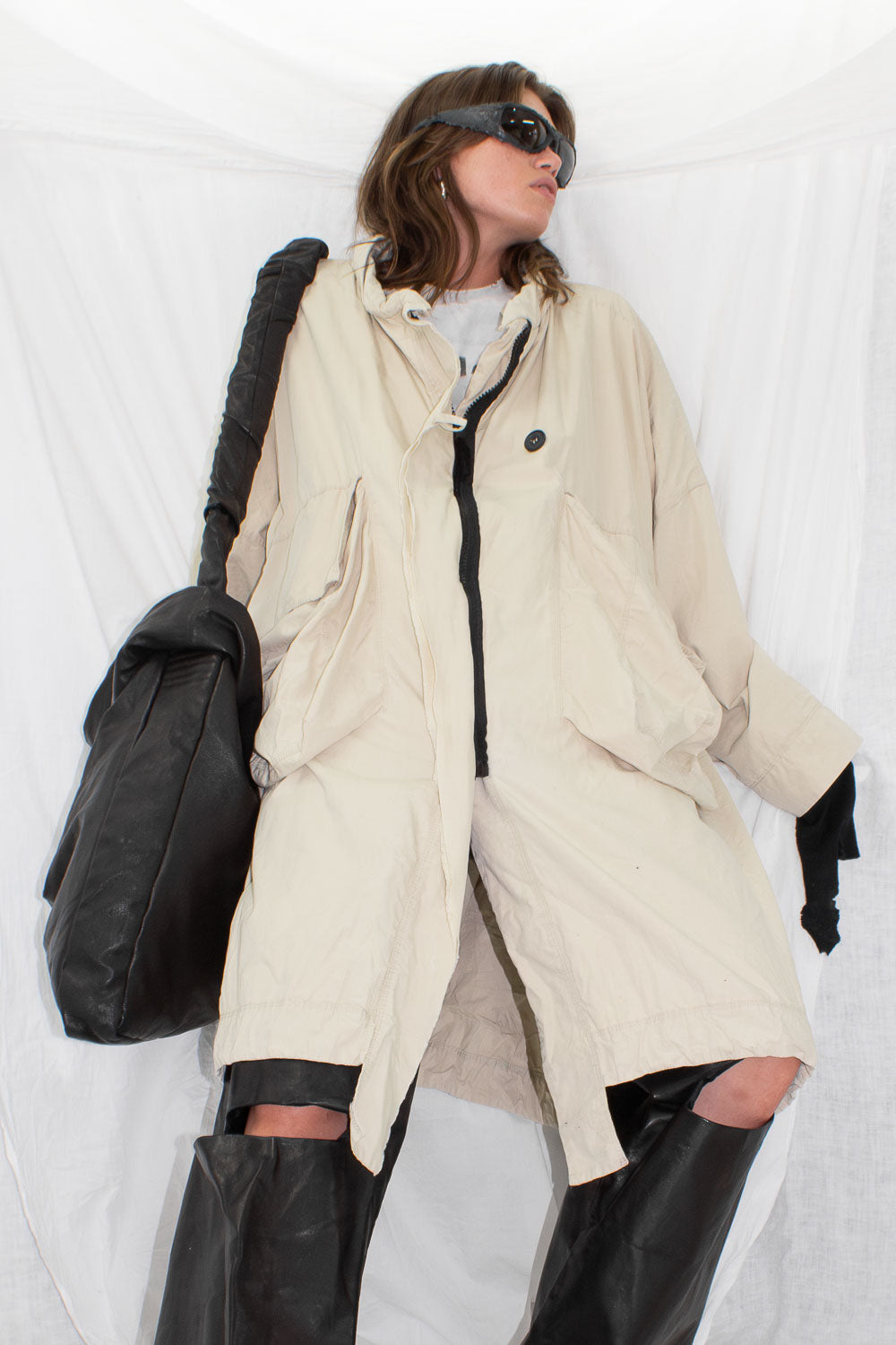 Model in beige parka with hood up, three-quarter angle showing draped pocket construction