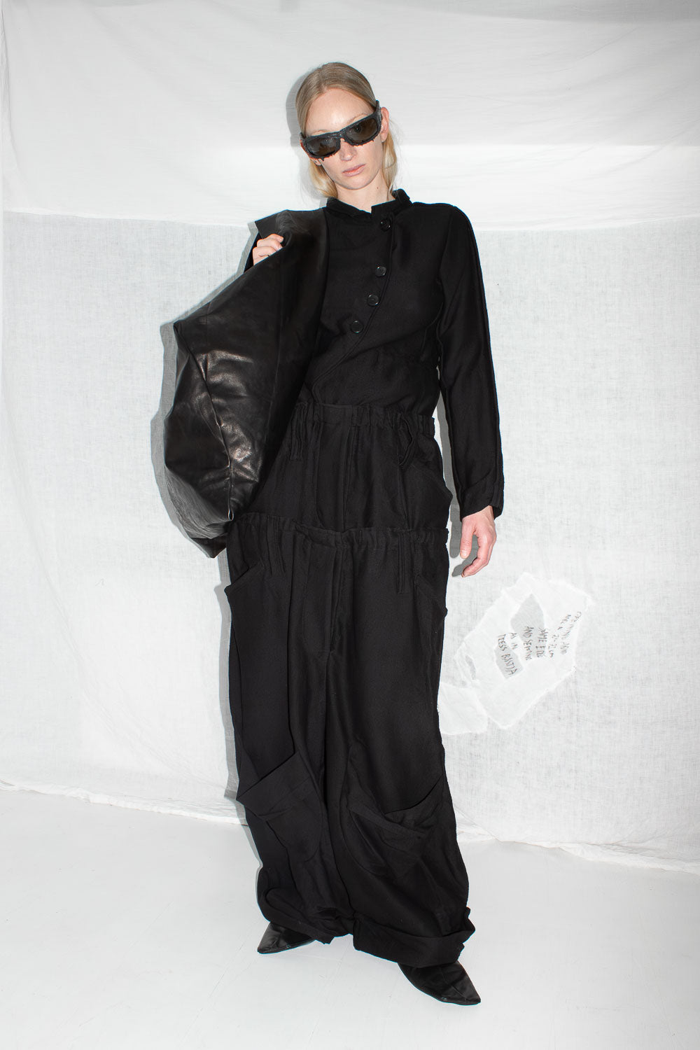 Model wearing black long-sleeve top with oversized trousers, carrying leather bag, full styling view